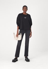 Black oversized sweatshirt with logo, dark jeans, and black platform shoes. Holding a light-colored bag with logo strap. Neutral background.