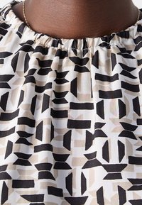 Gros plan d'une personne portant un collier et un chemisier blanc avec des formes géométriques noires et beiges et un col élastique.