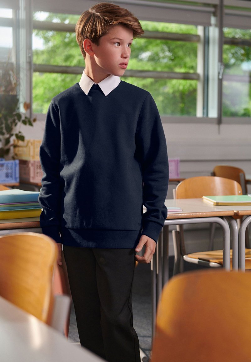 Tengerészeti kereknyakú pulóver fehér galléros inggel alatta, fekete nadrággal párosítva. Egy osztályteremben, fából készült padokkal.
