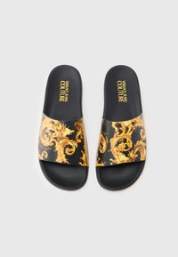 Chanclas negras con una correa ancha que presenta un patrón dorado ornamentado. Texto "Versace Jeans Couture" impreso en dorado en la correa. Suela de goma.