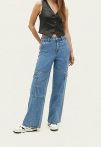 Wijd-uitlopende blauwe denim broek met meerdere zakken, een hoge taille en een nette afwerking. Gestoeld met een zwart mouwloos shirt en sneakers.