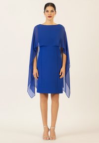 Blaues Kleid mit transparenten, fließenden Kapuzenärmeln. Glatter Stoff, knielanger Rock, runder Ausschnitt und minimale Details. Silberne Riemchensandalen.