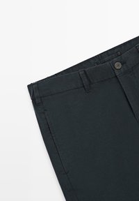 Donkergrijze broek plat gevouwen, met tailleband met knoop, lussen voor riem en zijzak tegen een witte achtergrond.