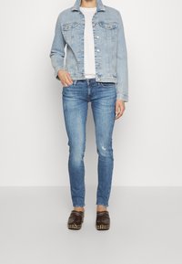 Chaqueta de mezclilla azul claro con cierre de botones, dos bolsillos en el pecho, combinada con jeans ajustados azul oscuro y zuecos marrones con tachuelas doradas.