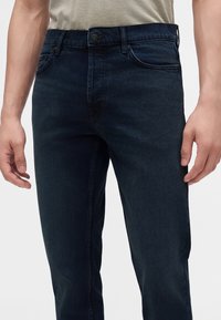 Homme portant un jean en denim bleu foncé et une chemise beige clair, les mains détendues le long du corps, sur un fond uni.