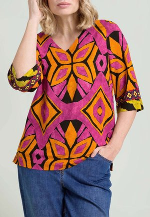 Donna che indossa una blusa con motivi vivaci rosa, arancioni e gialli a maniche tre quarti e jeans blu, con la mano in tasca.