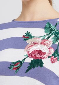 Primer plano de una camisa a rayas moradas y blancas con detallados bordados florales en rosa y verde en el pecho cerca del escote.