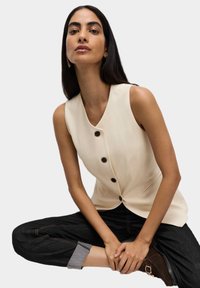 Cremefarvet ærmeløs vest med v-hals og mørke knapper, parret med mørk denim bukser med optrækkede manchetter og kontrastsyet.