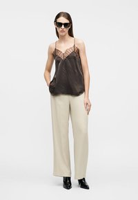 Γυναίκα που φοράει μαύρο τοπ με δαντελένια διακόσμηση, μπεζ παντελόνι με φαρδιά πόδια, μαύρες μυτερές μπότες και μαύρα γυαλιά ηλίου, στέκεται μπροστά σε ένα απλό λευκό φόντο.