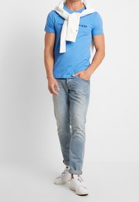 Muž v světle modrém tričku Tommy Hilfiger, světlých džínách, bílých teniskách a bílém svetru přes ramena.