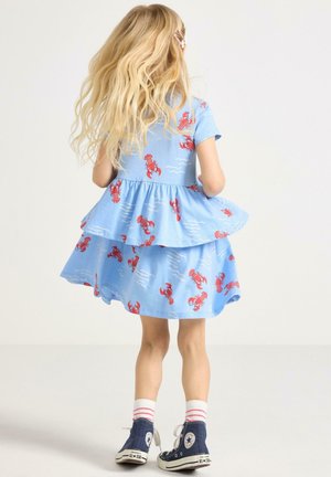 Blyhåret barn set bagfra iført en blå kjole med røde hummelprint, hvide sokker med røde striber og marineblå højt opsnitne sneakers.