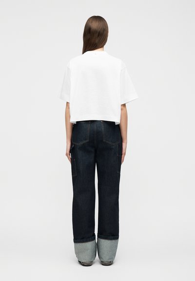 Γυναίκα που φαίνεται από πίσω φορώντας λευκό κοντό μπλουζάκι και σκούρο τζιν με φαρδιές παντελόνες και μεγάλες μανσέτες, στέκεται μπροστά σε απλό ανοιχτόχρωμο φόντο.