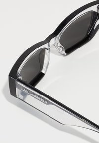 Lunettes de soleil surdimensionnées noires et transparentes avec un design géométrique, des montures noires épaisses, des verres sombres et un logo gravé sur la charnière de la branche.