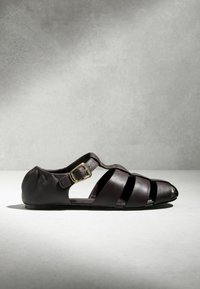 Sandale pour homme en cuir marron foncé de style pêcheur, avec une lanière à boucle dorée, semelle plate et design à bout ouvert sur une surface claire.