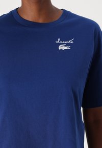 Tamsiai mėlyna medvilninė marškinėliai su apvalia apykakle, su baltu siuvinėtu "Lacoste" tekstu ir logotipu krūtinėje. Trumpomis rankovėmis, glotnus paviršius.