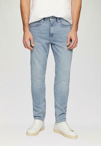 Jean en denim bleu clair avec une coupe slim, doté de cinq poches, d'un léger effet décoloré et de détails de couture. Porté avec des baskets blanches.