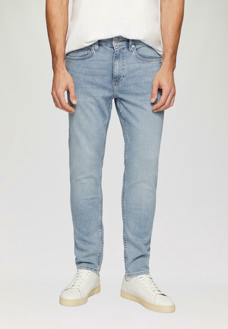 Jean en denim bleu clair avec une coupe slim, doté de cinq poches, d'un léger effet décoloré et de détails de couture. Porté avec des baskets blanches.