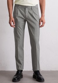 Mann, der hellgraue maßgeschneiderte Hosen und schwarze Leder-Schnürschuhe trägt, steht vor einer weißen strukturierten Wand.