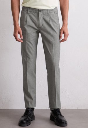 Man in een lichtgrijze, op maat gemaakte broek en zwarte leren veterschoenen, staand tegen een witte, gestructureerde muur.