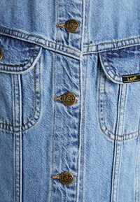 Lichtblauwe spijkerjacket met gouden Lee knopen, dubbele borstzakken en zichtbaar stikwerk. Klassiek ontwerp, casual uitstraling.