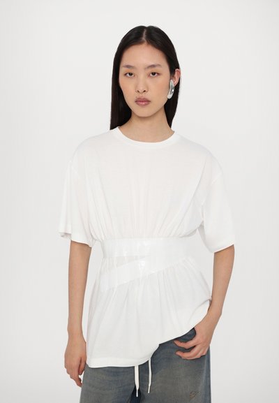 Vit bomullst-shirt i oversized passform med en blank, figurnära midjedetalj; har rund hals och korta ärmar, i kombination med denimbyxor.