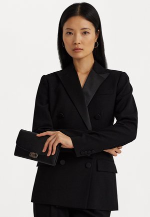 Femme aux longs cheveux noirs portant un blazer noir à double boutonnage tenant une pochette noire avec un logo doré sur fond blanc.