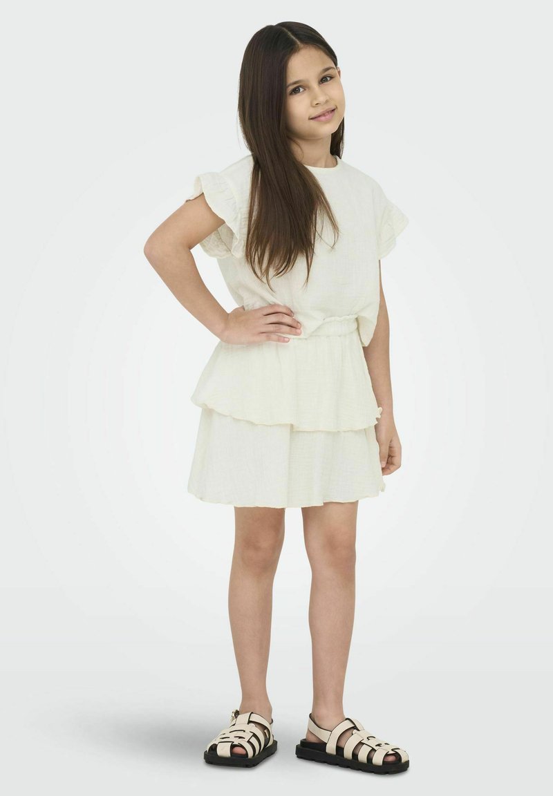 Biała, warstwowa sukienka z falbanistymi rękawami, wykonana z lekkiego materiału. Noszona z czarnymi płaskimi sandałami o paskowej konstrukcji. Dziecko stoi pewnie.