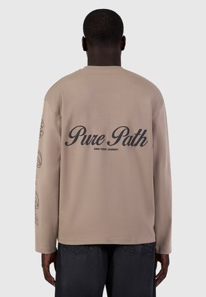 Person wearing beige long-sleeve shirt with "Pure Path Own Your Journey" text on back and dark pants, shown from behind.