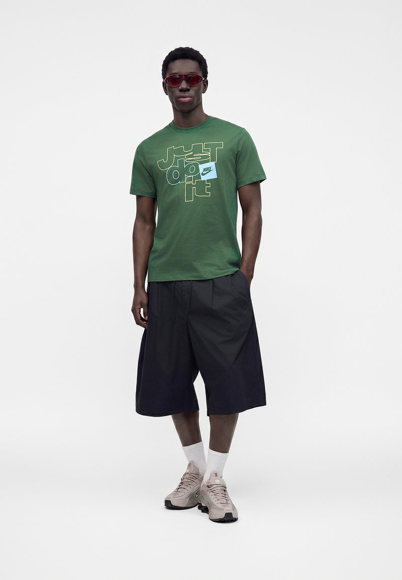Mand iført røde solbriller, grøn Nike T-shirt, sorte vide shorts, hvide sokker og beige sneakers, står med den ene hånd i lommen.