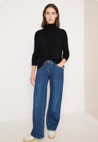 Pull en tricot noir avec texture côtelée, col montant ; associé à un jean large bleu. Le mannequin porte des baskets blanches. Fond de couleur unie.