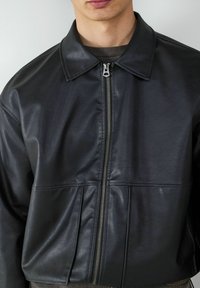 Giubbotto in pelle nera con colletto, chiusura con zip frontale e dettagli a pieghe nell'orlo. Texture liscia e forma strutturata.