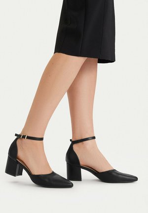 Donna che indossa scarpe nere con tacco medio, punta a punta e cinturini alla caviglia, in piedi con le gambe incrociate, vestita con una gonna nera.