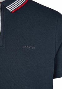 Polo shirt bleu marine en tissu texturé, avec un col rayé en blanc, gris et rouge, et un logo en relief subtil au centre.