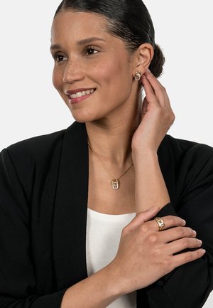 Goldkette mit einem verlinkten Anhänger, passender goldener Ring, getragen mit einem schwarzen Blazer über einem weißen Oberteil. Glatte Texturen und polierte Oberflächen.