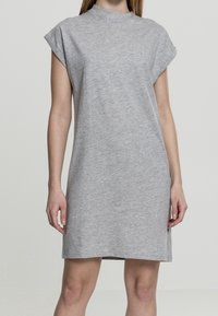 Femme portant une robe décontractée simple, à manches courtes, de longueur genou, de couleur gris clair, se tenant contre un fond uni et clair.