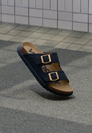 Sandale slide noire avec deux sangles ajustables et boucles dorées, semelle intérieure en liège et semelle en caoutchouc sur l'asphalte avec un fond carrelé.