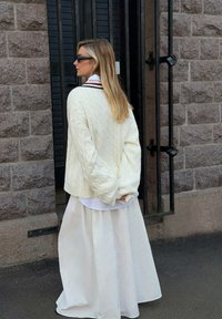 Suéter de punto de cable crema usado sobre una camisa blanca con cuello y una falda blanca fluida, con el cabello largo y rubio peinado suelto.