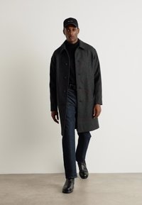 Mørk grå overfrakk laget av wool-blend, med knappfront, sidelommer og krage, brukt over svart turtleneck og jeans.