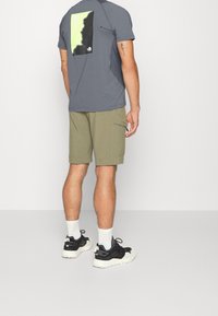 Šedé tričko s krátkým rukávem a grafikou na zadní straně, doplněné světle olivovými šortkami. Na nohou jsou černé sportovní boty a bílé ponožky.