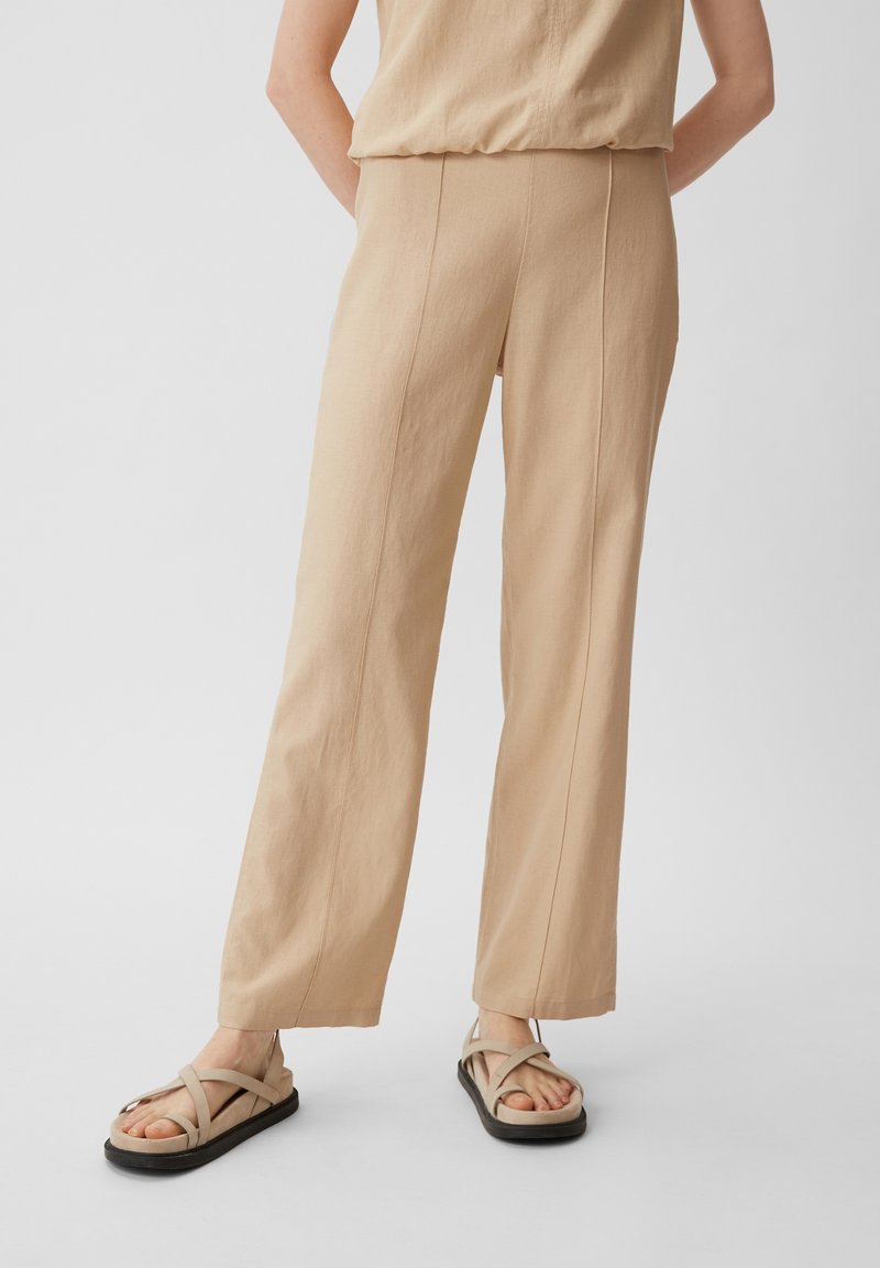 Beige, weit geschnittene Hose aus strukturiertem Material mit vertikalen Nähten. Kombiniert mit beigen Sandalen mit Kreuzriemen auf einer schwarzen Sohle.