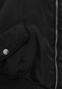 Chaqueta bomber negra con un exterior de nailon brillante, cierre de cremallera, puños elásticos y un bolsillo con botón a presión en el lado izquierdo.