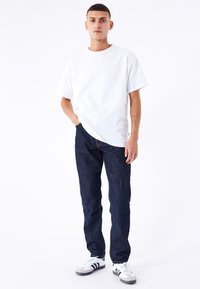Camiseta blanca de manga corta, jeans de denim azul y zapatillas en blanco y negro. La figura está de pie con las manos en los bolsillos frente a un fondo blanco.