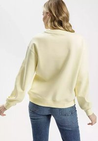 Żółta, oversize'owa bluza z długimi rękawami, ściągaczami przy nadgarstkach i okrągłym dekoltem, w połączeniu z niebieskimi dżinsami. Miękka struktura.