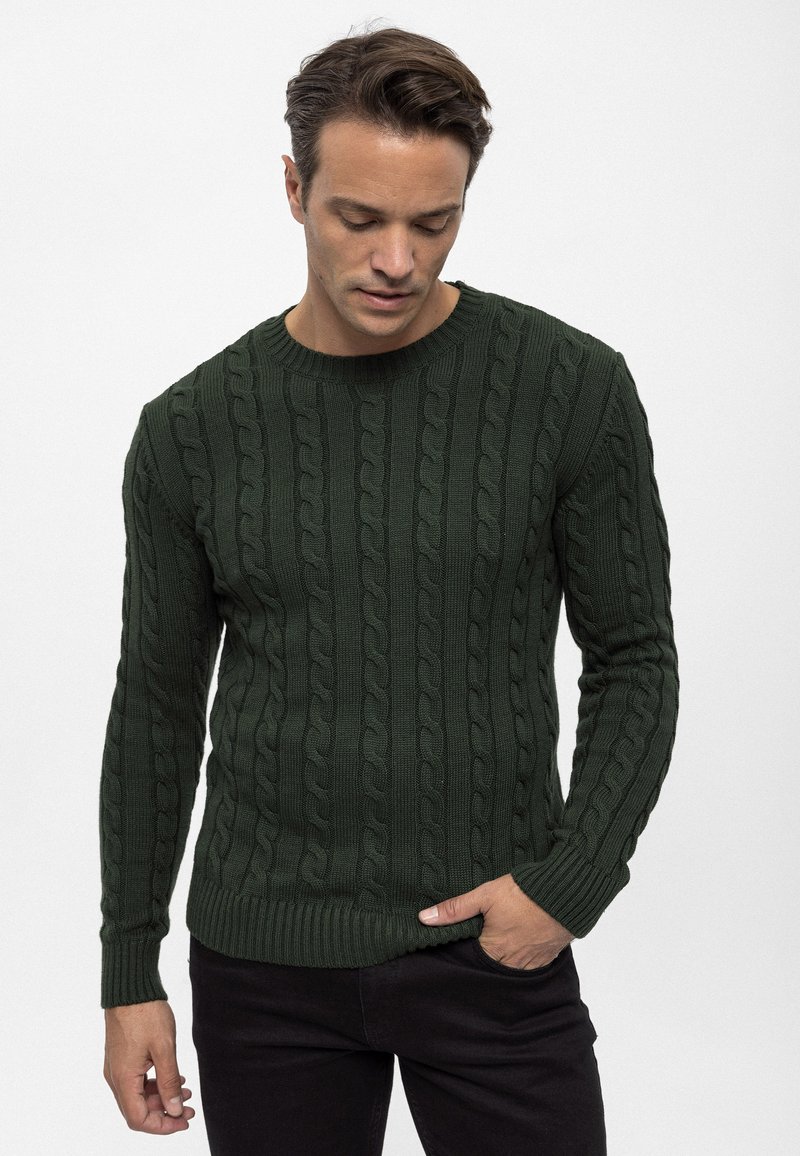 Maglione verde lavorato a maglia con motivo a trecce, collo rotondo e polsini a costine. Indossato con jeans scuri che mostrano un dettaglio di tessitura.
