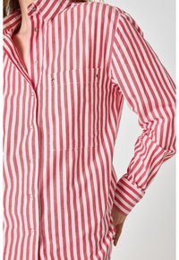 Camicia a righe rosse e bianche con colletto, maniche lunghe e taschino sul petto con dettagli in metallo argentato.