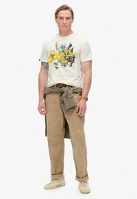 Camiseta gráfica beige con estampado colorido y mangas cortas, combinada con pantalones chinos marrón claro y deportivas crema. Suéter atado en la cintura.