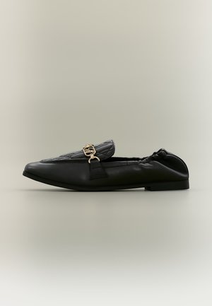 Schwarzer flacher Lederschuh mit gerafftem Rückenteil, gesteppter Oberseite und goldfarbenem Metallschmuckverschluss auf beigem Hintergrund.