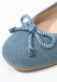 Denimballerina i blått med en texturerad yta. Har en tvinnad blå satinrosett vid tåspetsen och en ljus färgad sula.