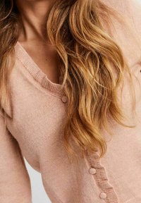 Frau mit langen welligen blonden Haaren, die eine hellrosa, geknöpfte Strickjacke mit V-Ausschnitt trägt.