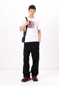 Tricou alb cu text grafic negru și roșu, asociat cu pantaloni negri de tip cargo și adidași negri cu accente roșii. Ține în mână o geantă mică neagră.